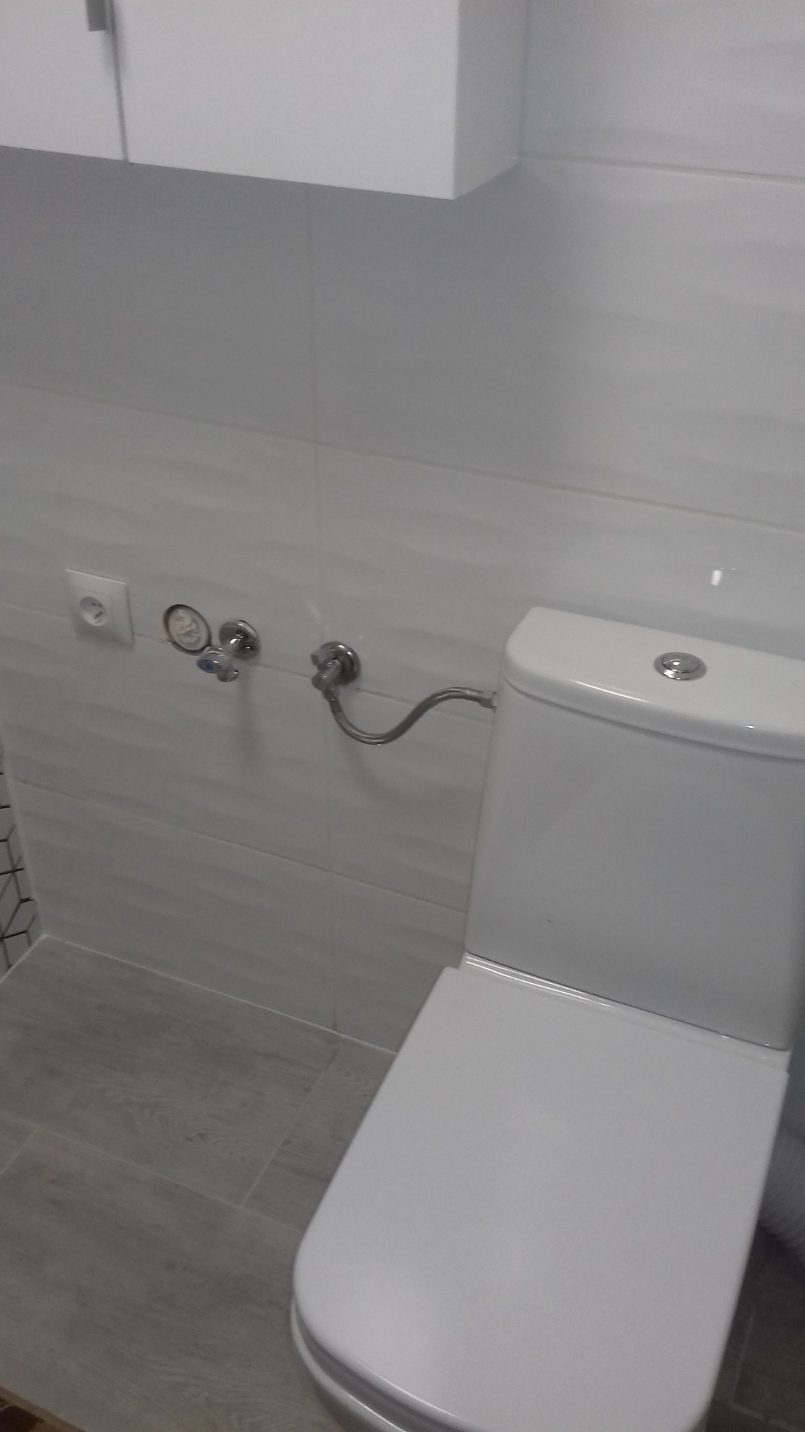 Biała toaleta z podłączonymi rurami i zaworami na ścianie z jasnoszarymi płytkami, widoczne gniazdko elektryczne i fragment szafki wiszącej.