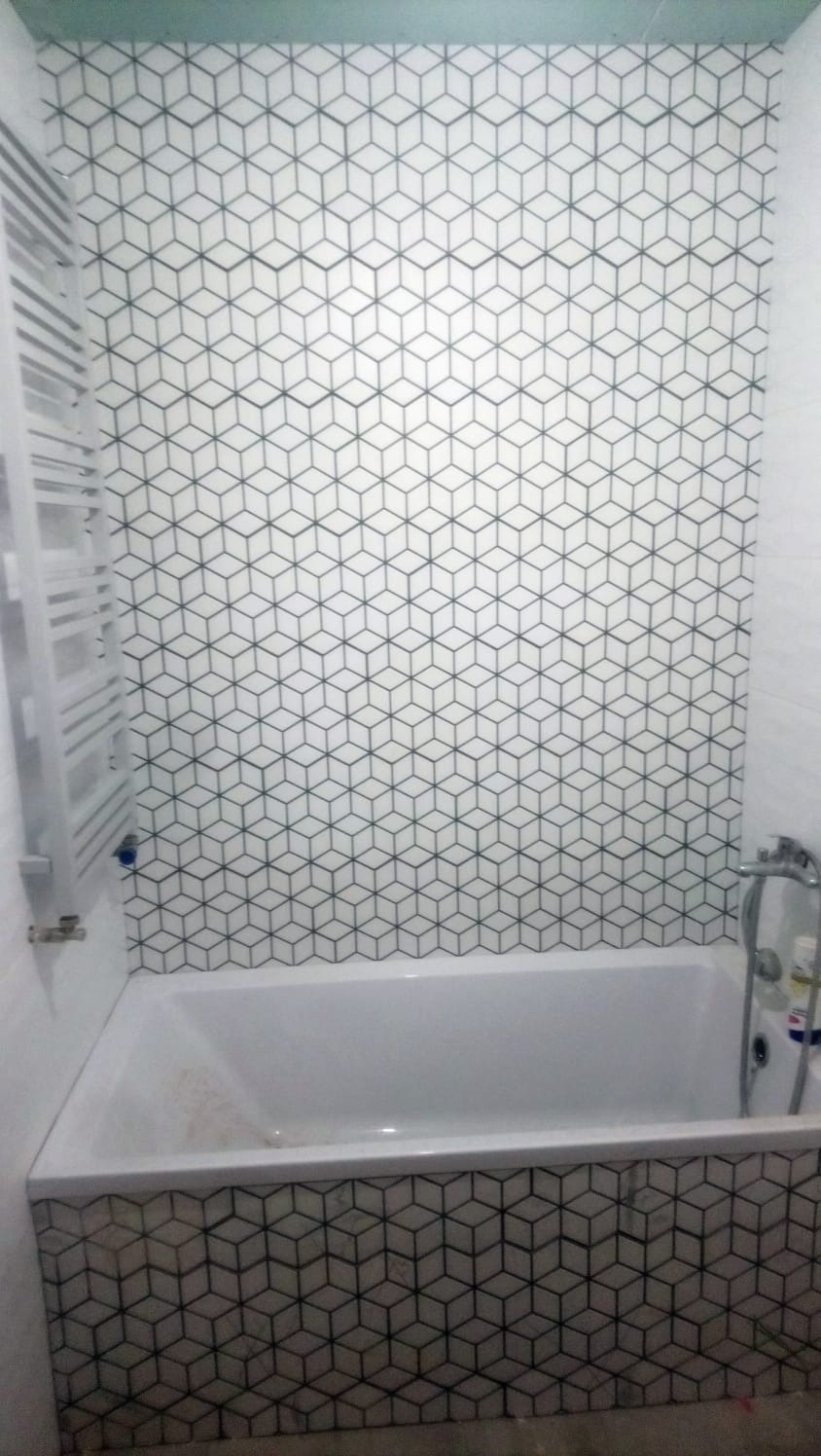 Wanna obłożona mozaiką z geometrycznym wzorem kostek 3D, podobny wzór na ścianie, grzejnik drabinkowy po lewej, bateria wannowa po prawej stronie.