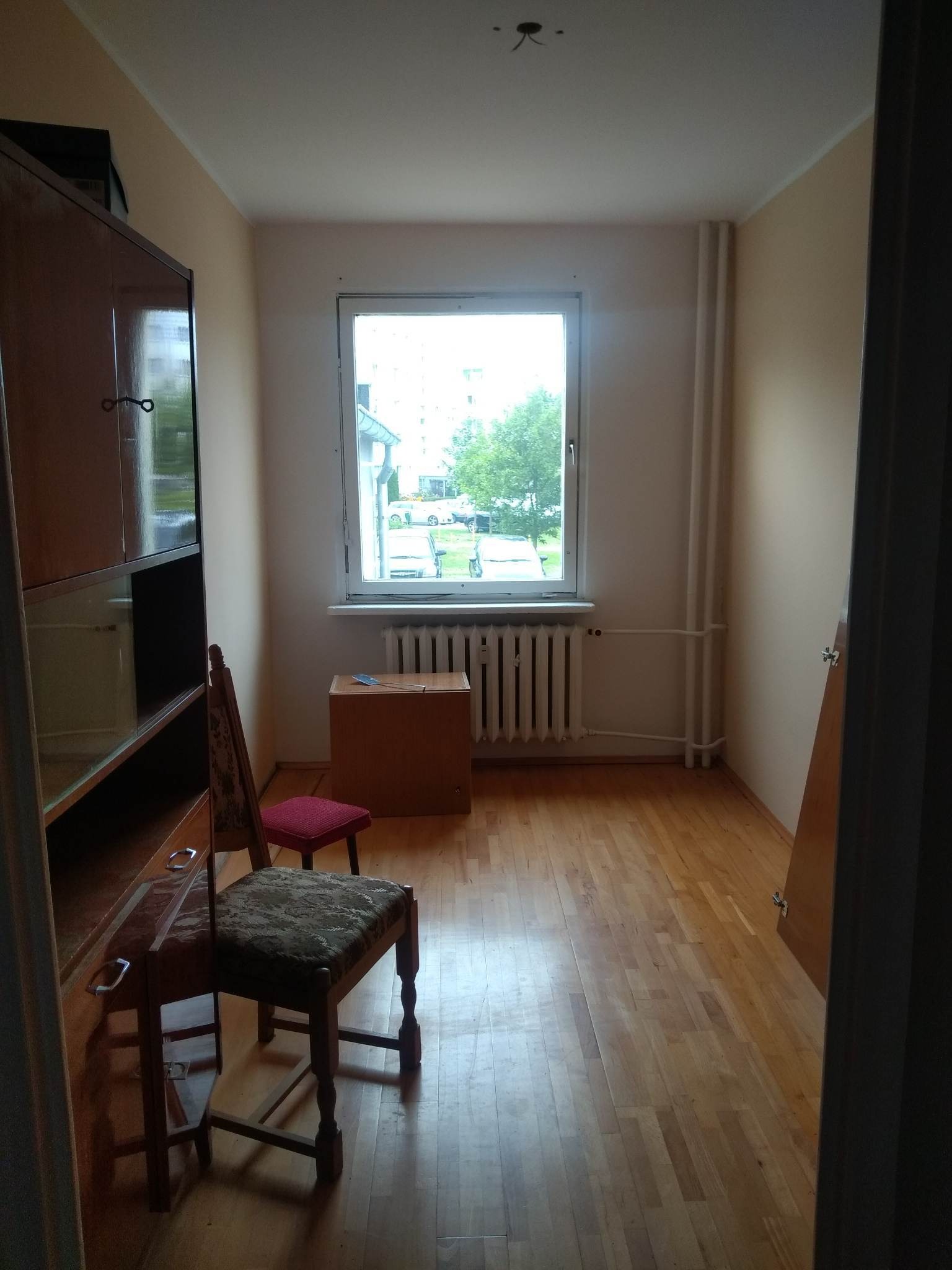 Pusty pokój z parkietem, grzejnikiem pod oknem i starymi meblami przed remontem. Widoczne ślady po instalacjach na suficie.