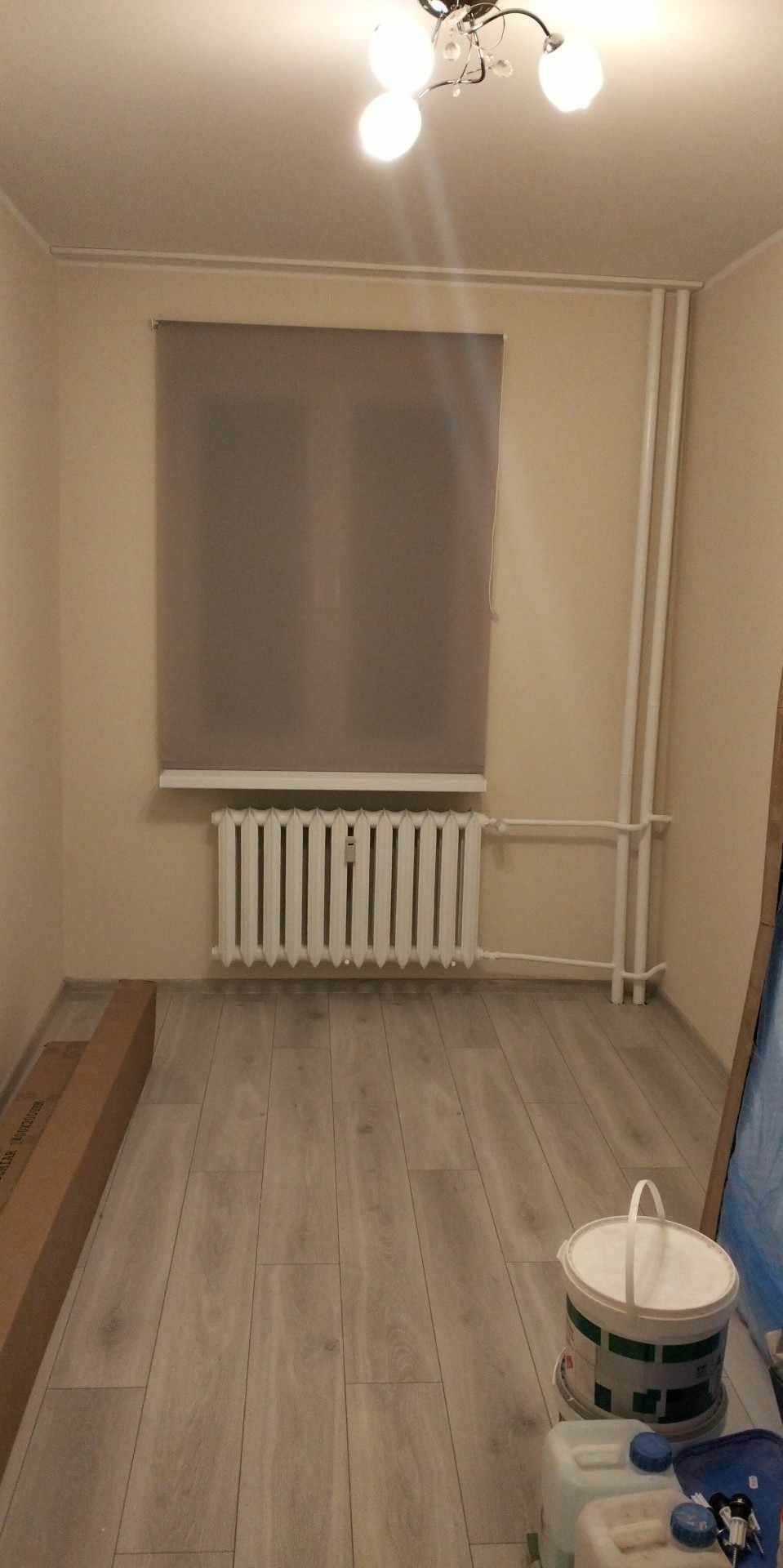 Pusty pokój po remoncie z nową podłogą imitującą drewno, zasłoniętym oknem roletą w kolorze szarym, białym grzejnikiem i niedokończonymi pracami w rogu, gdzie widać pojemniki z farbą i akcesoria.