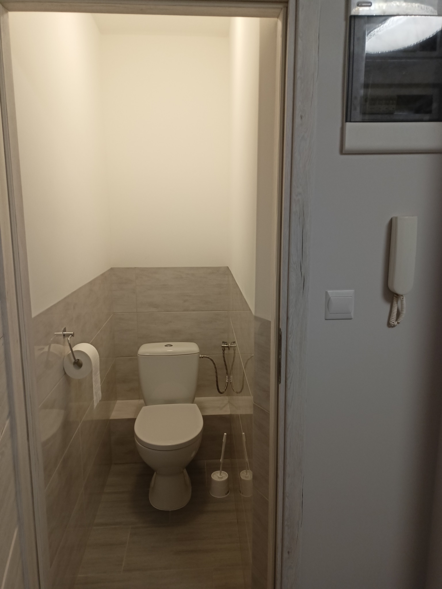 Wykończona toaleta w odcieniach szarości, z widoczną zabudową z płytek ceramicznych do połowy wysokości ścian, wiszący podajnik na papier toaletowy i szczotka do WC w rogu, widok zza otwartych drzwi.