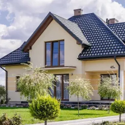 Nowoczesny dom jednorodzinny z ciemnym dachem z metalowych paneli na tle błękitnego nieba z chmurami, widoczne rynny i numer domu 45A.