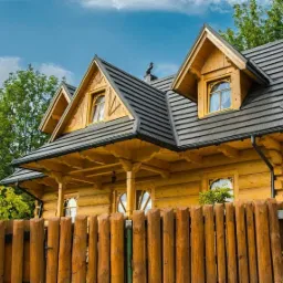 Drewniany dom w stylu góralskim z ciemną, nowoczesną dachówką, widok od frontu z drewnianym płotem, błękitne niebo w tle.