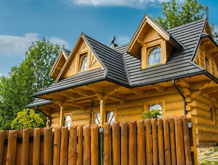 Drewniany dom w stylu góralskim z ciemną, nowoczesną dachówką, widok od frontu z drewnianym płotem, błękitne niebo w tle.
