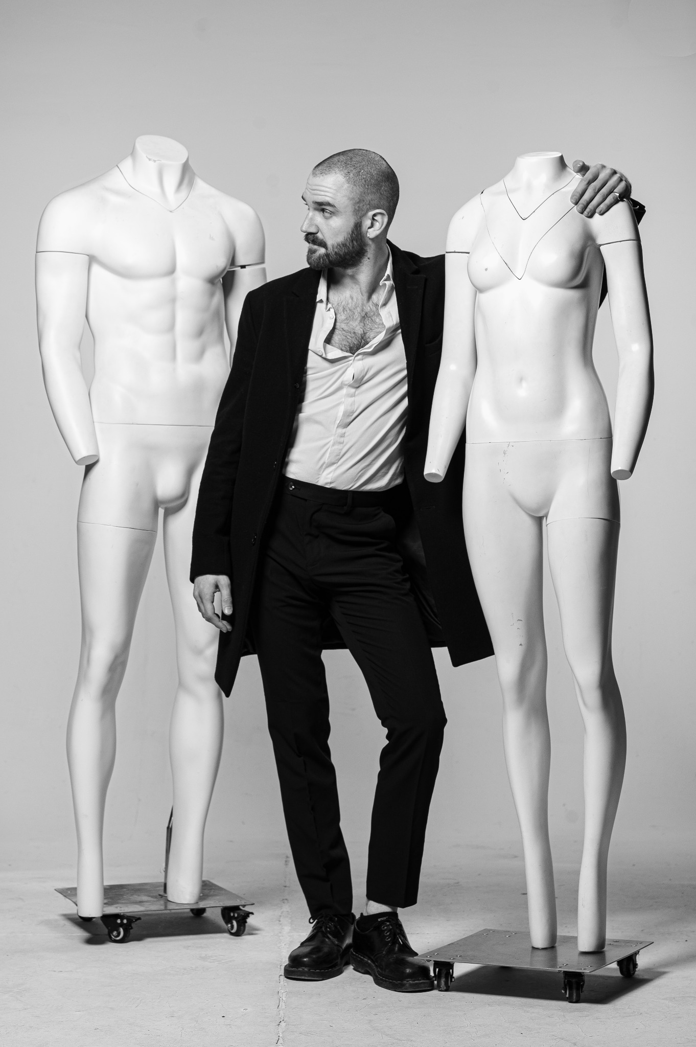 Mężczyzna w rozpiętej koszuli i płaszczu, obejmujący ramię manekina na kółkach, drugi manekin obok, czarno-białe zdjęcie studyjne.