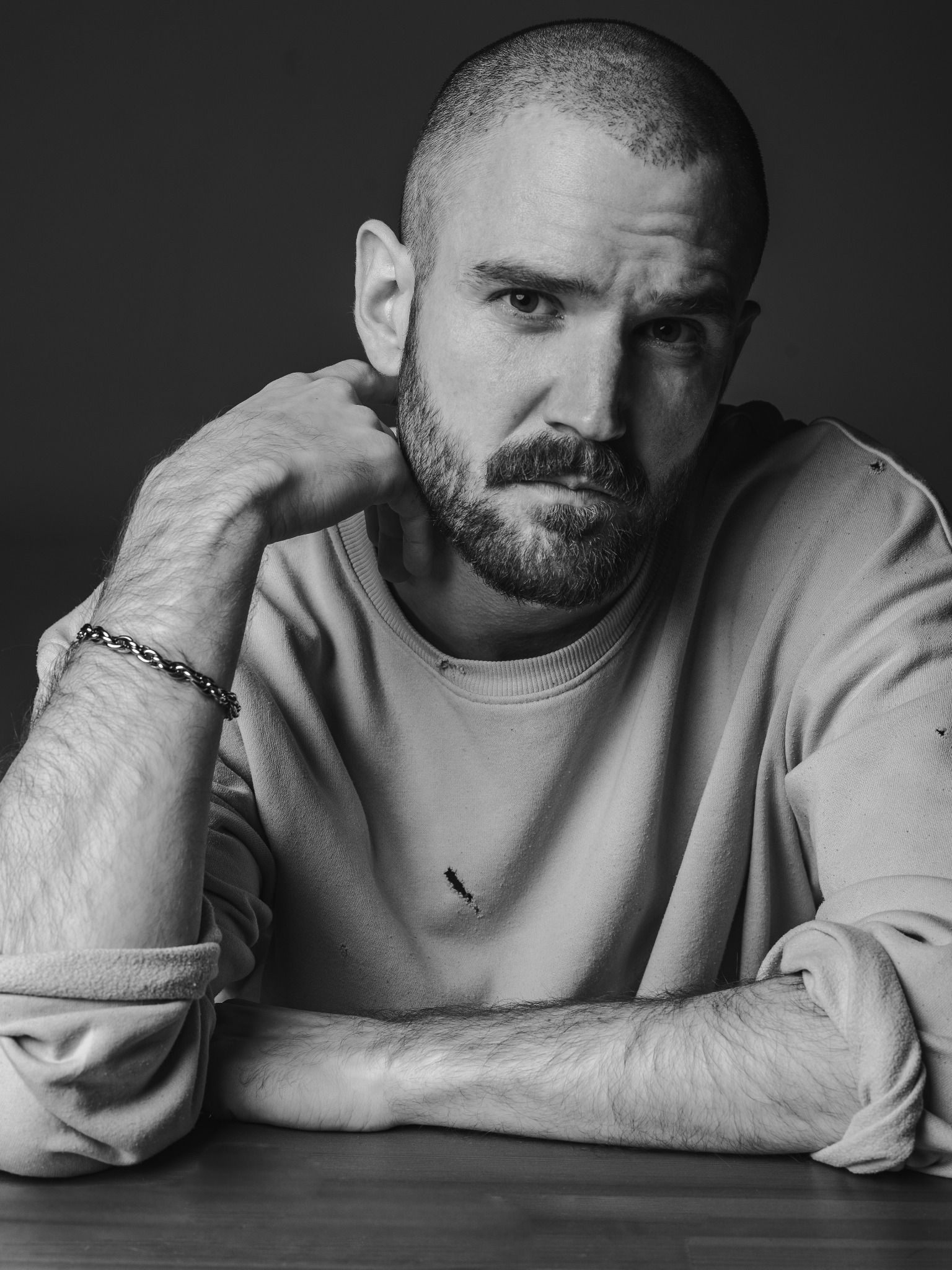 Portret mężczyzny z brodą, opierającego brodę na dłoni, z bransoletką na nadgarstku, w jasnej bluzie z drobnymi uszkodzeniami, na ciemnym tle.