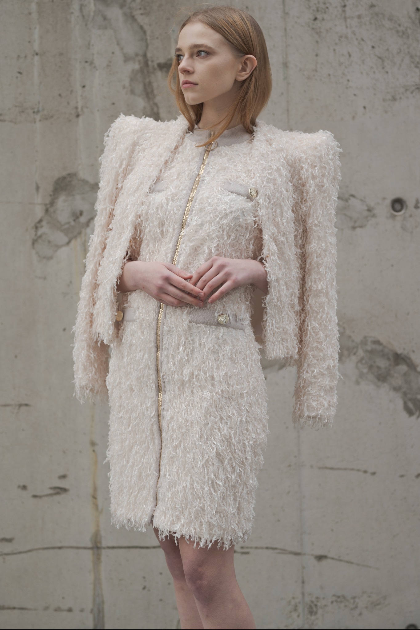 Kobieta pozuje w kremowej sukience i marynarce z frędzlami na tle surowej, betonowej ściany. Sukienka zapinana na złoty zamek, marynarka luźno zarzucona na ramiona.