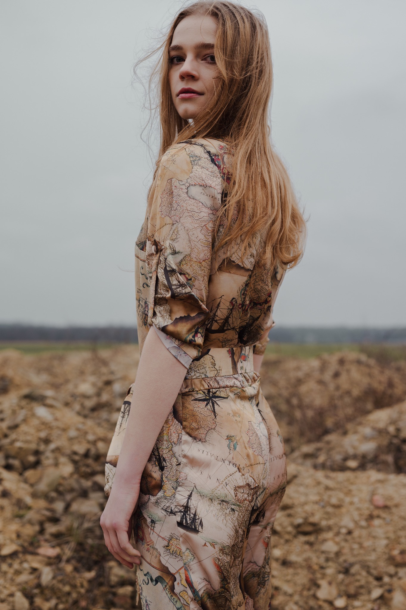 Młoda kobieta spogląda przez ramię, ubrana w satynowy kombinezon z motywem mapy świata, na tle zaoranego pola i pochmurnego nieba.