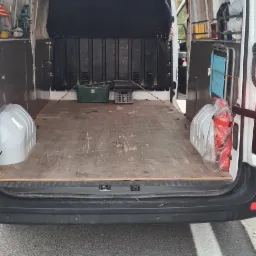 Wnętrze białego busa z drewnianą podłogą, widoczne dwa zielone pojemniki, gaśnica oraz metalowy wózek z niebieskim siedziskiem, oświetlenie wewnętrzne, otwarte tylne drzwi.