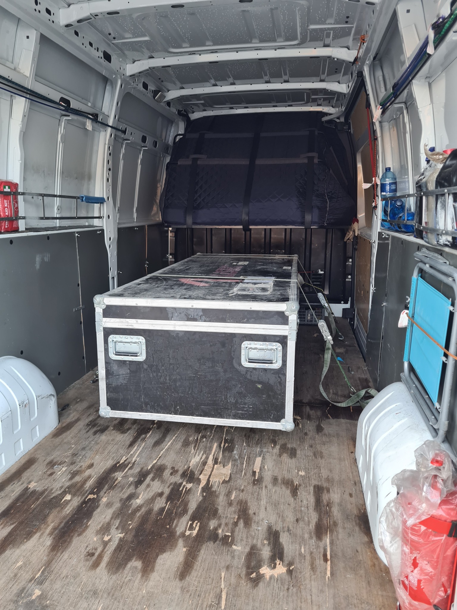 Wnętrze białego busa z zabezpieczonym, czarnym skrzyniowym ładunkiem, dodatkowo zabezpieczonym pasami transportowymi, widoczna apteczka, butelki z wodą i krzesło turystyczne.