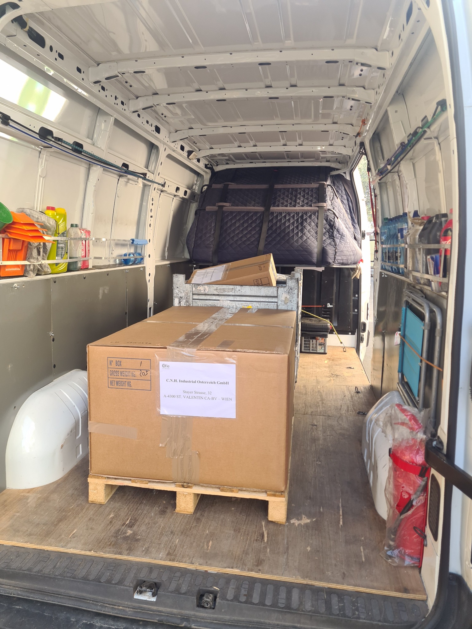 Wnętrze białego busa załadowane kartonami na paletach, zabezpieczonymi kocami, z widocznymi butelkami wody i gaśnicą po bokach.