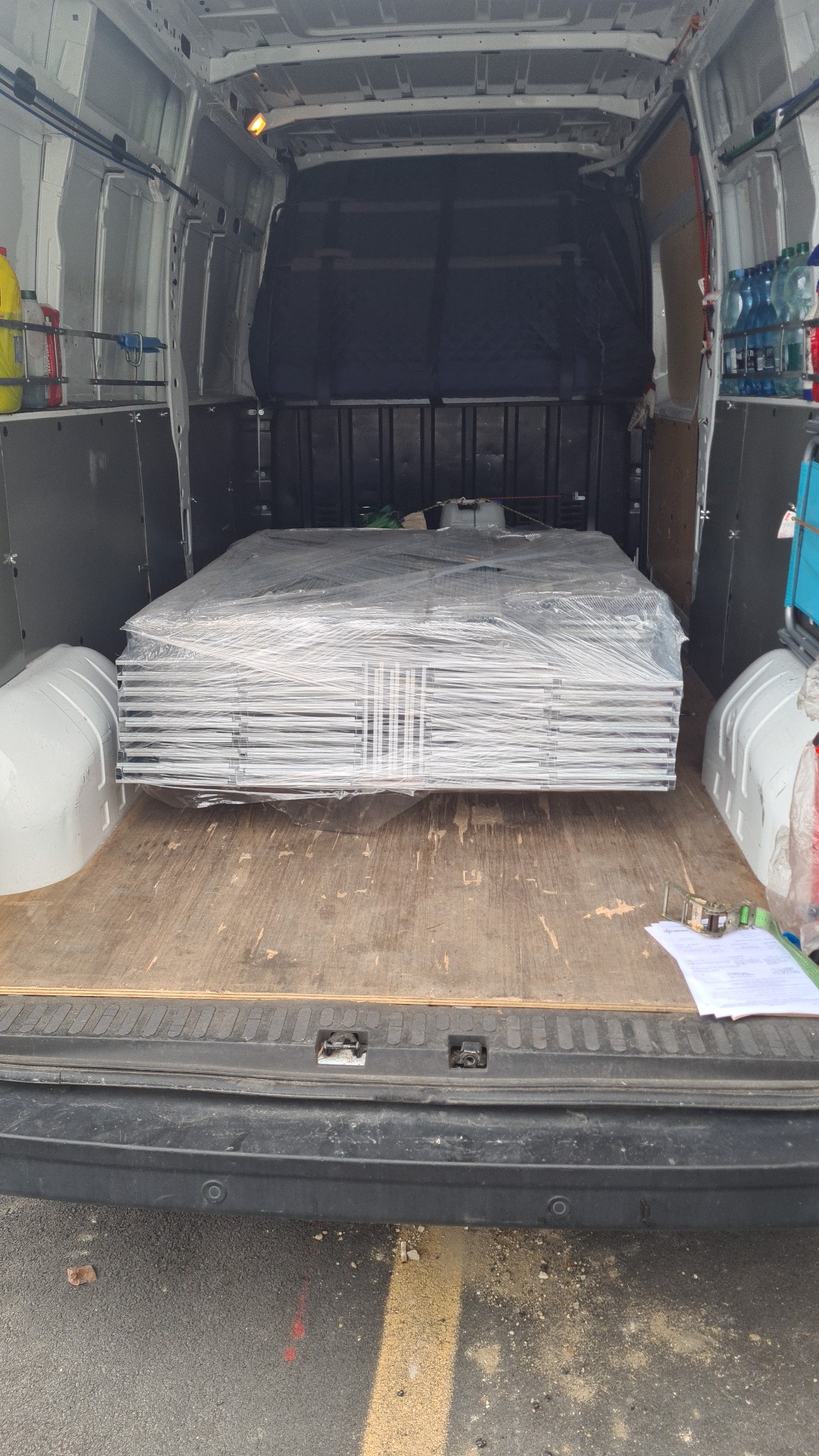 Wnętrze busa z zabezpieczonym stosem paneli lub płyt owiniętych folią, gotowych do transportu, widoczne butelki z wodą i płyny na półkach po bokach.