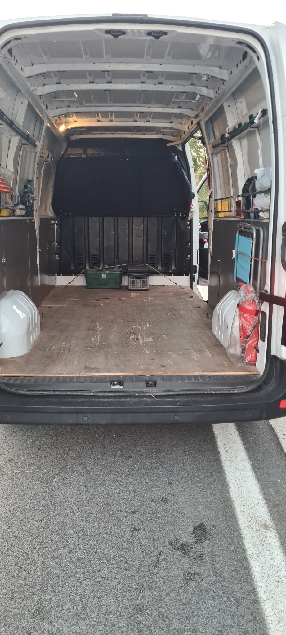 Wnętrze białego busa z drewnianą podłogą, widoczne dwa zielone pojemniki, gaśnica oraz metalowy wózek z niebieskim siedziskiem, oświetlenie wewnętrzne, otwarte tylne drzwi.