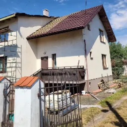 Dom jednorodzinny w trakcie wymiany dachu, częściowo pokryty brązową blachodachówką, widoczny ruszt drewniany i rusztowanie, materiały budowlane za żelazną bramą.