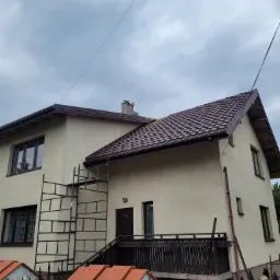 Dwupiętrowy dom z kremową elewacją i nowym brązowym dachem z blachodachówki, widoczne rusztowanie przy ścianie oraz fragment starego dachu z dachówki ceramicznej w tle, pochmurne niebo.