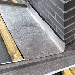 Dach-Błysk - Szczegółowe ujęcie obróbki blacharskiej komina na dachu, z widoczną konstrukcją drewnianą i czarną membraną dachową, wykonane w pochmurny dzień.