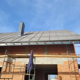 Budowa domu w trakcie krycia dachu szarą dachówką, widoczny komin z szarej cegły, rusztowania i niebieskie ubranie wiszące na rusztowaniu.