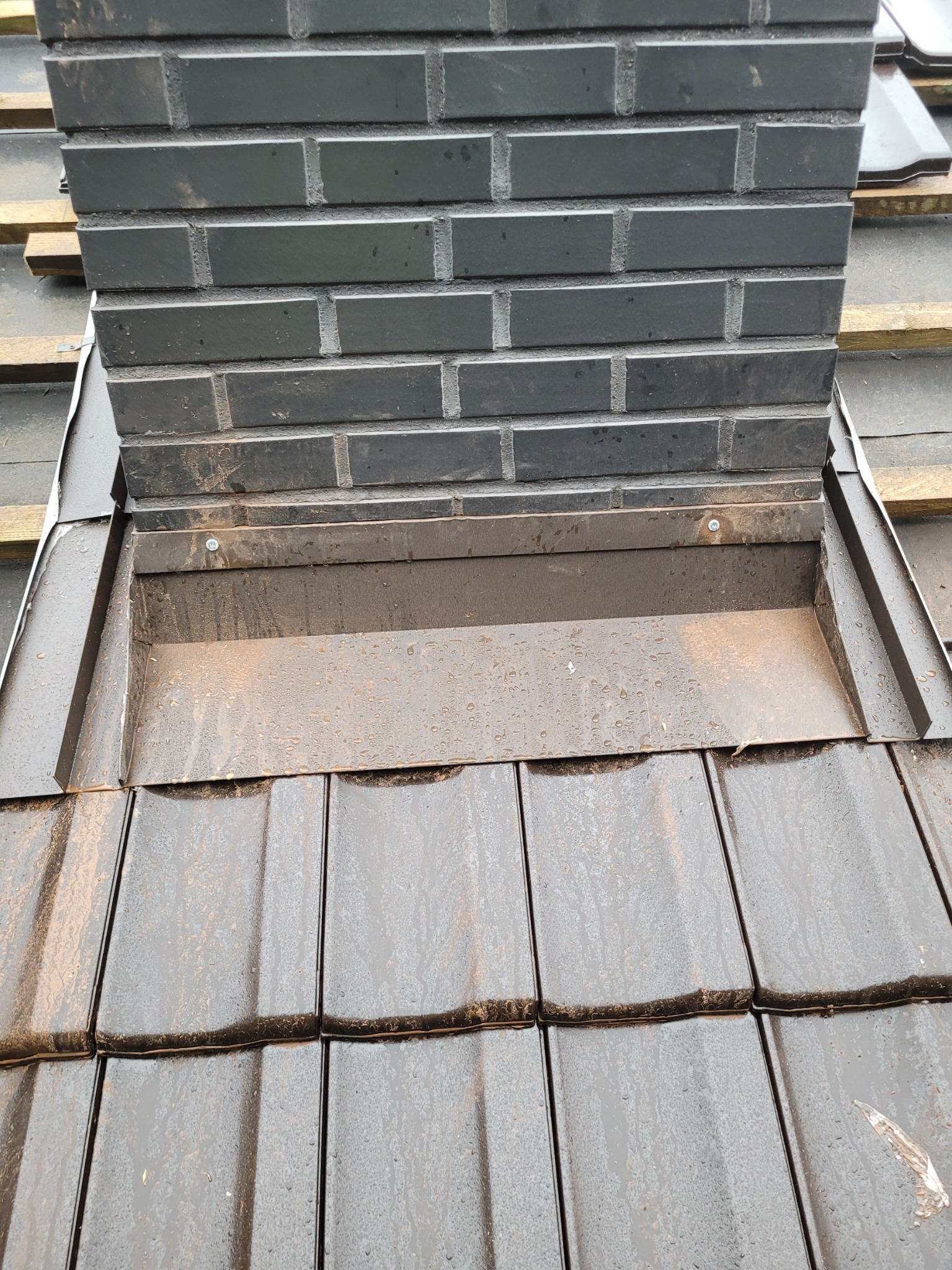 Szczegółowe ujęcie czarnego komina z cegły klinkierowej, połączonego z dachem pokrytym ciemnymi dachówkami, z widoczną obróbką blacharską i śladami wilgoci.