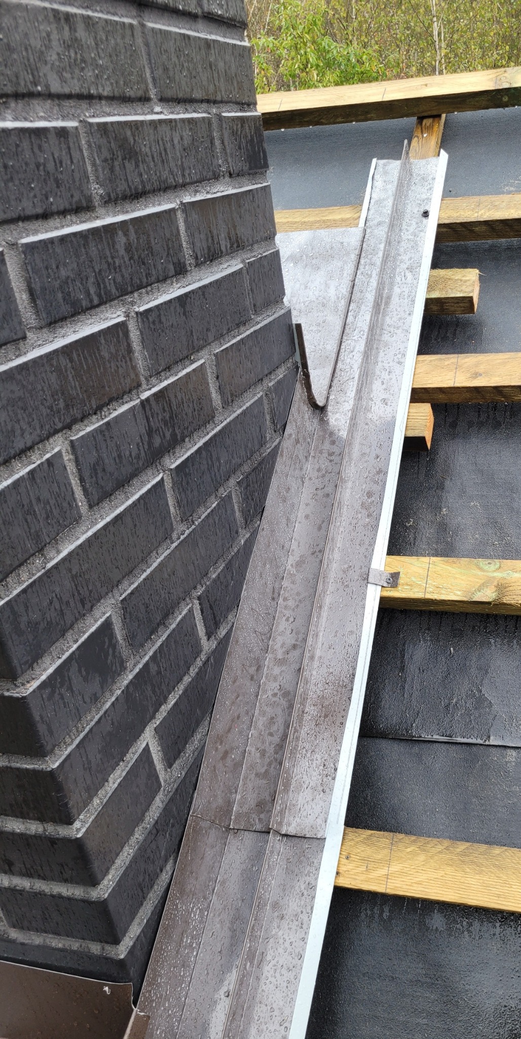 Mokry, ciemny komin z cegły klinkierowej sąsiaduje z nowo zamontowaną, metalową rynną koszową na tle czarnego pokrycia dachowego i drewnianej konstrukcji dachu.