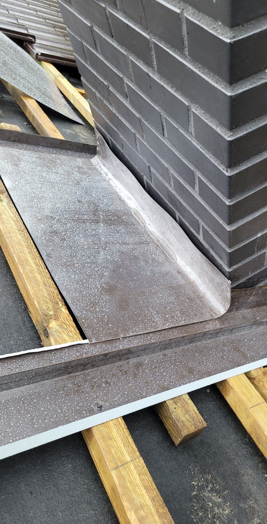 Szczegółowe ujęcie obróbki blacharskiej komina na dachu, z widoczną konstrukcją drewnianą i czarną membraną dachową, wykonane w pochmurny dzień.