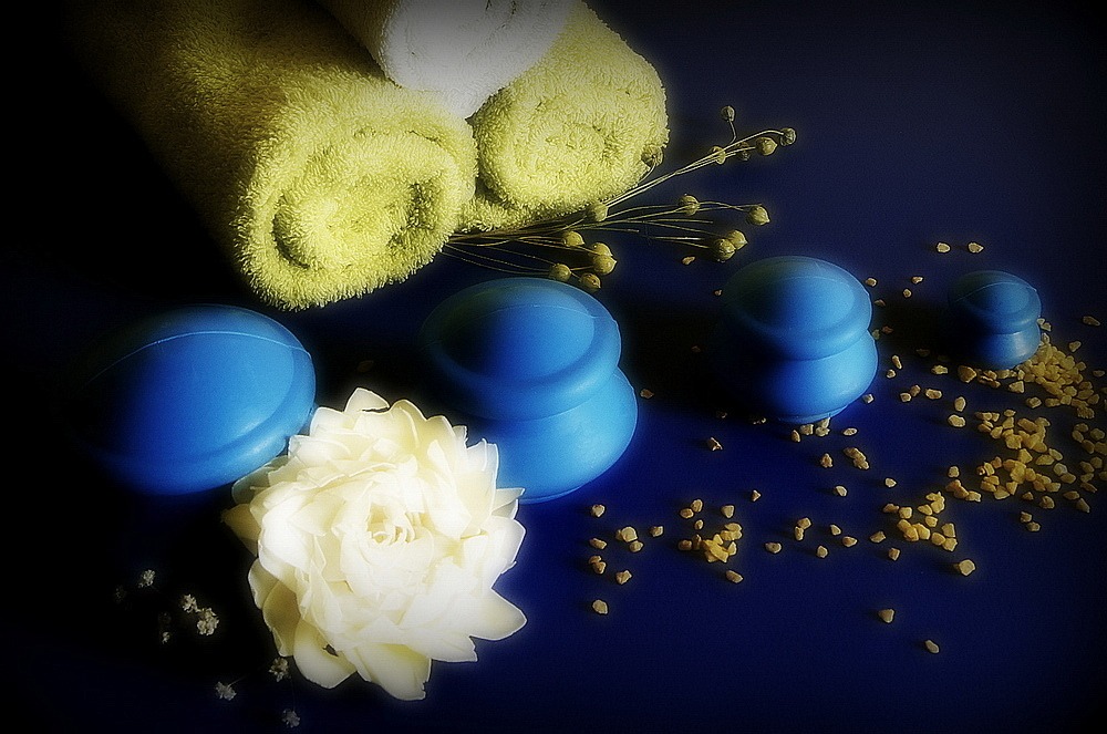 Martwa natura z niebieskimi bańkami do masażu próżniowego, zwiniętymi zielonymi ręcznikami, białym kwiatem i rozsypanymi kryształkami soli na ciemnoniebieskim tle.