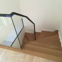 Drewniane schody spiralne z czarną, szklaną balustradą, widok z góry, białe ściany i podłoga w jasnym odcieniu drewna.