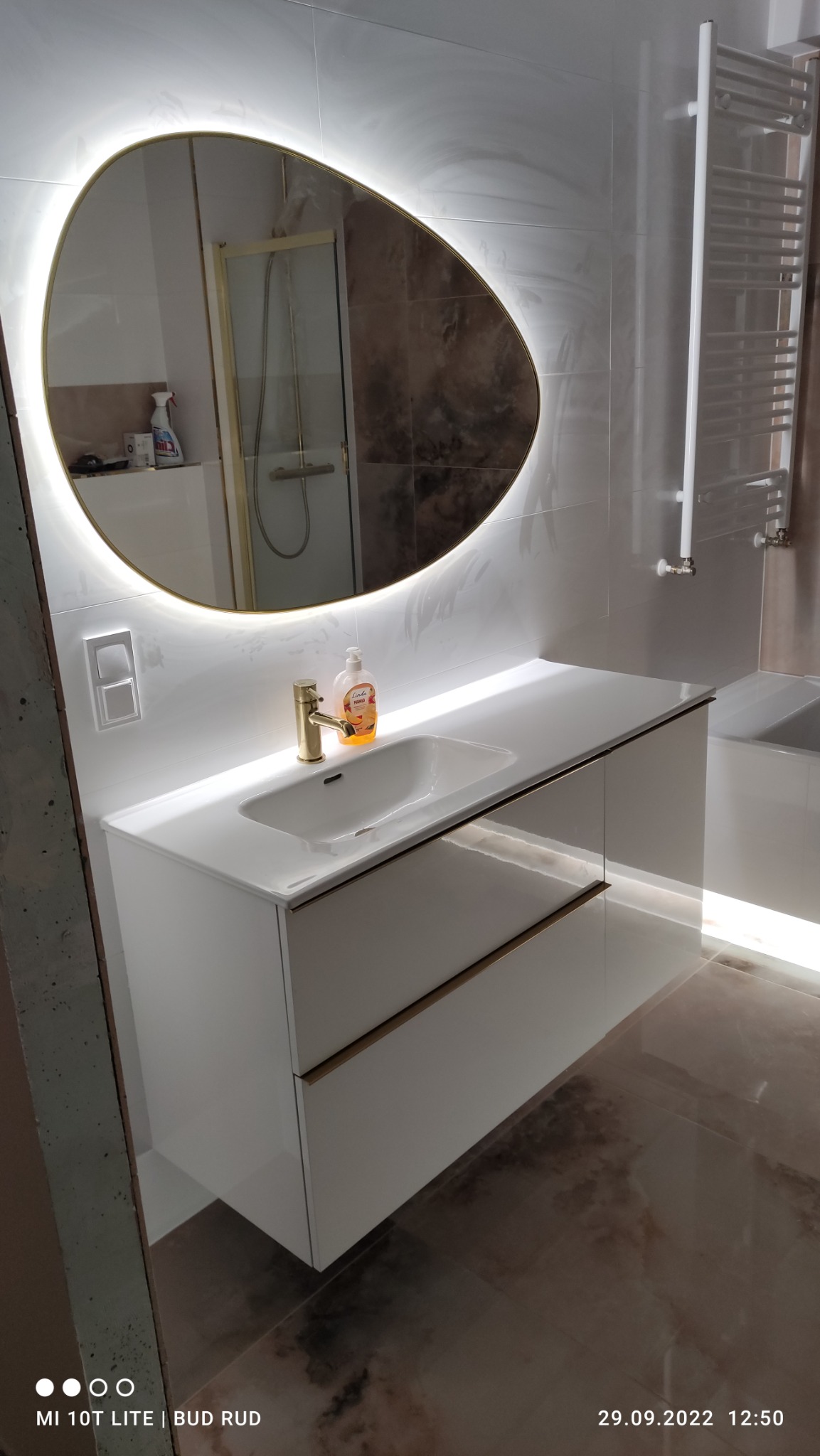 Nowoczesna łazienka z owalnym lustrem podświetlanym LED, białą szafką pod umywalką z złotymi akcentami i złotą baterią, brązowe płytki podłogowe i białe płytki ścienne.