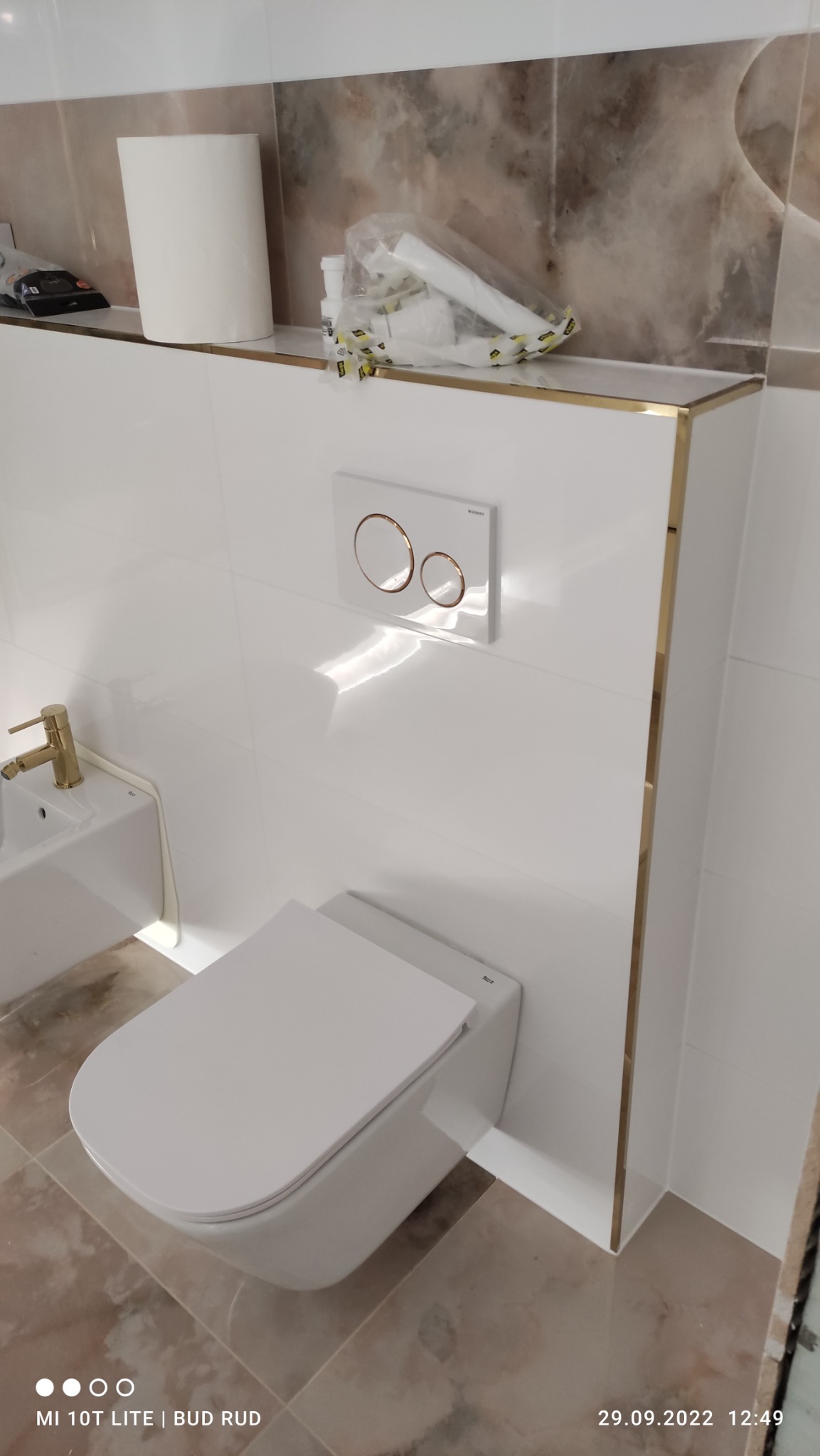 Nowoczesna, biała toaleta podwieszana z przyciskiem spłukującym w kolorze białym z miedzianymi obwódkami, zamontowana na tle białych płytek z marmurowym akcentem na górze i złotymi wykończeniami.