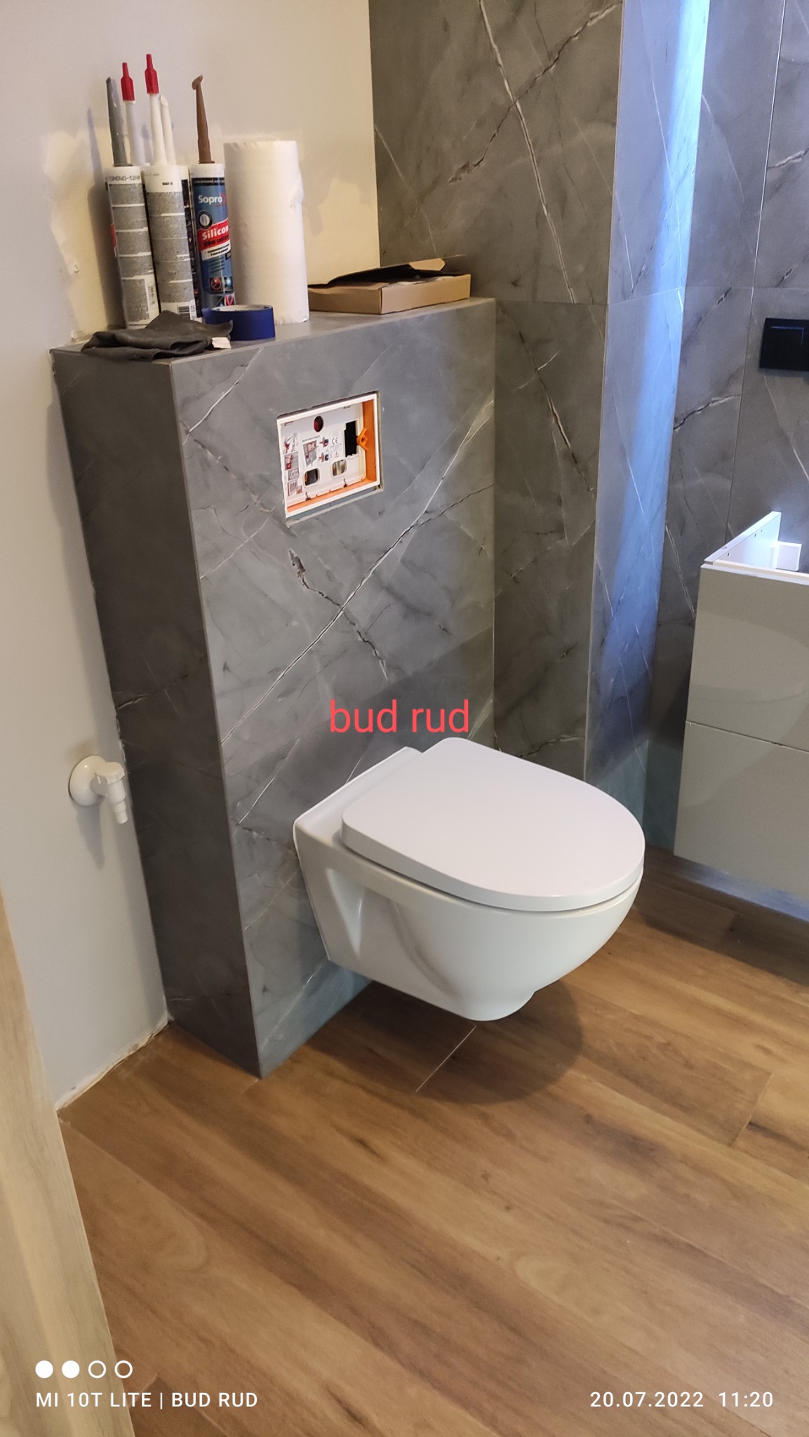 Podwieszana miska WC zamontowana na stelażu obudowanym płytkami imitującymi marmur, widoczne akcesoria montażowe i silikony na obudowie stelaża, drewnopodobna podłoga.