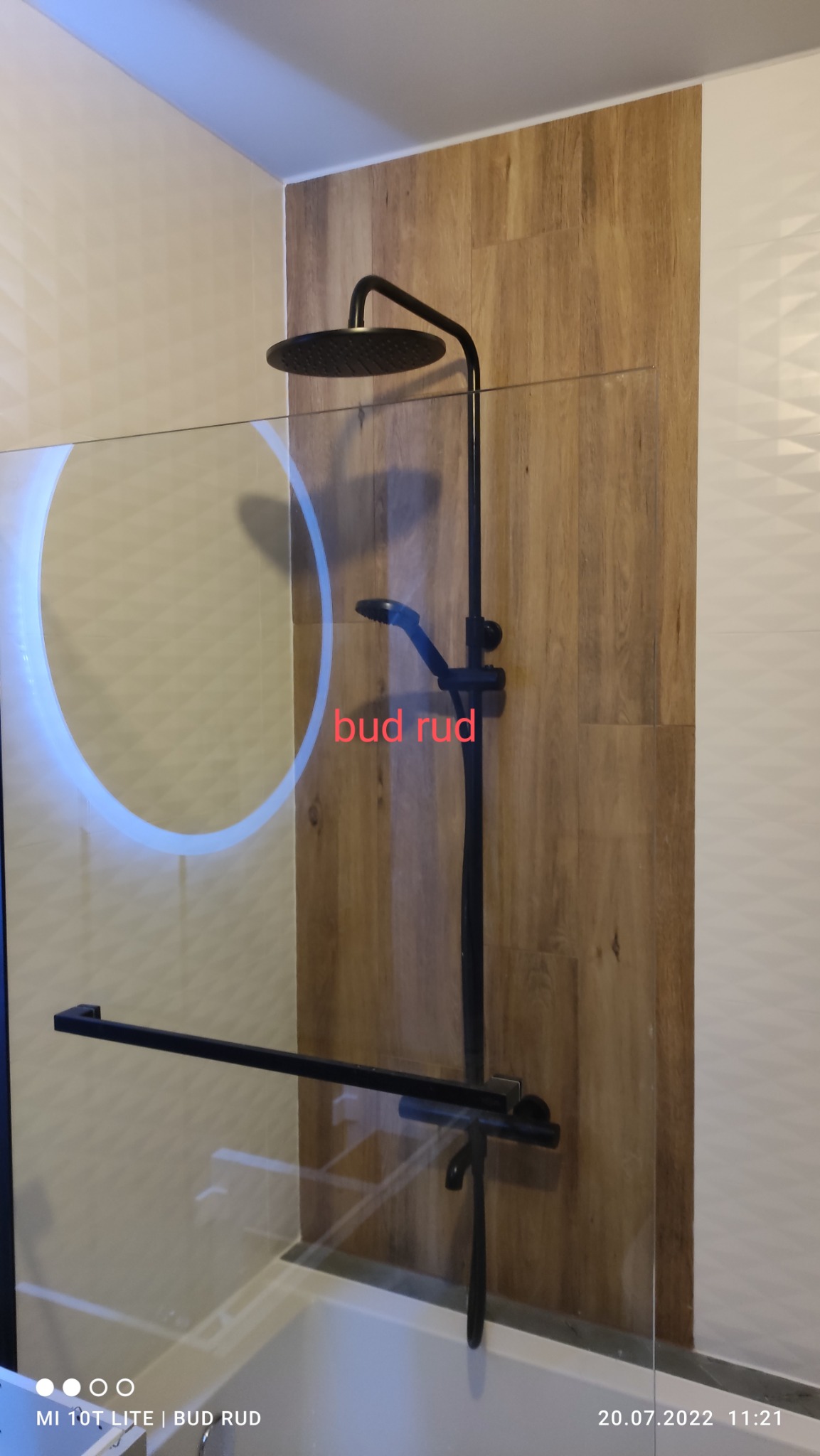 Nowoczesna kabina prysznicowa z czarną armaturą na tle płytek imitujących drewno i lustrem z podświetleniem LED, widok z boku