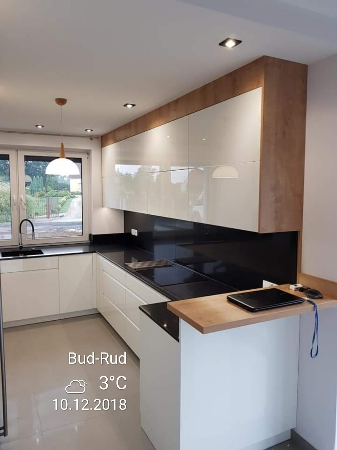 Nowoczesna kuchnia we Wrocławiu z białymi, lakierowanymi szafkami, czarnym blatem i drewnianymi akcentami, oświetlona punktowymi lampami sufitowymi i wiszącą lampą nad oknem.