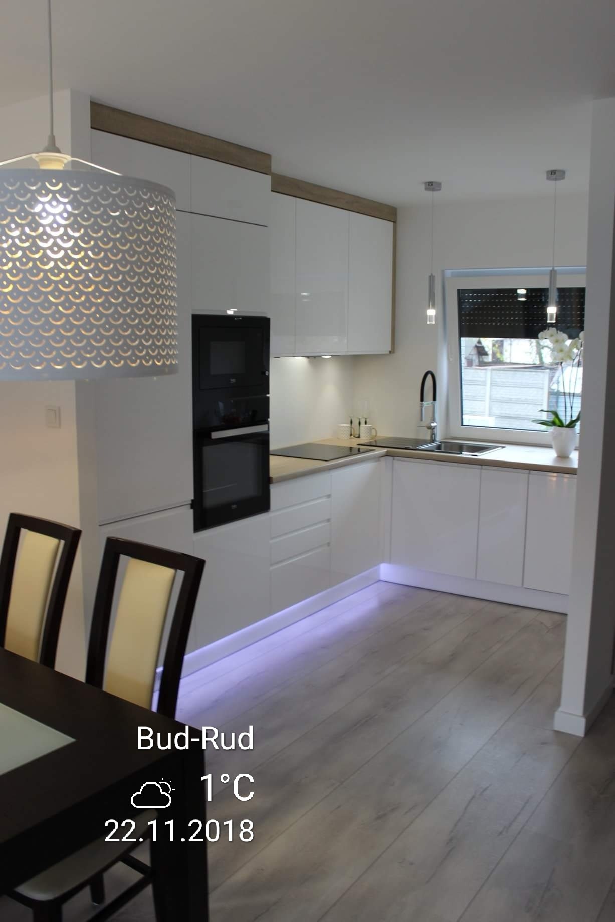 Jasna kuchnia w stylu skandynawskim z białymi szafkami, drewnianymi akcentami i podświetleniem LED u dołu, widok z perspektywy jadalni z ciemnym stołem i krzesłami.