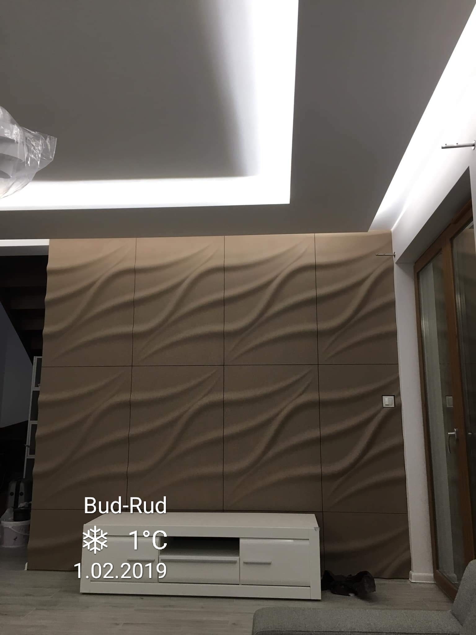 Ściana wykończona panelami 3D w odcieniach beżu, podświetlona listwą LED na suficie, z widoczną białą szafką RTV i fragmentem okna po prawej stronie. Na panelach widoczny delikatny wzór fal.