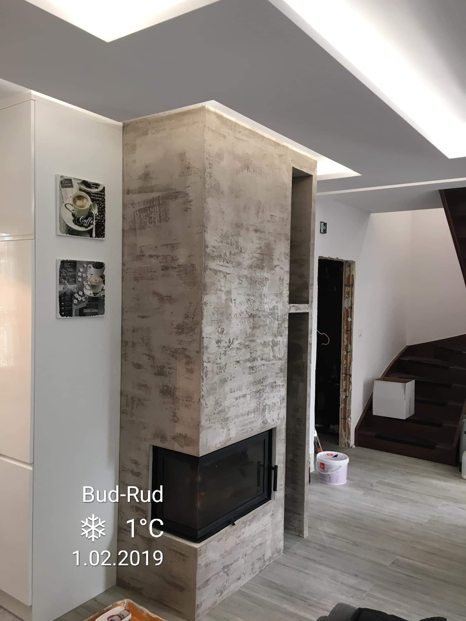 Nowoczesny kominek obłożony płytami imitującymi beton w jasnym salonie z białymi meblami i schodami w tle, widoczne dekoracje ścienne z motywem kawy.