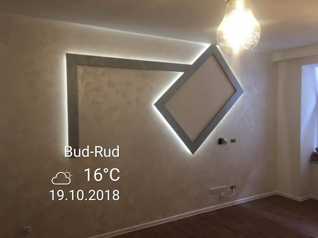 Ściana z dekoracyjną, podświetlaną ramą w kształcie rombu, jasna żarówka w górnym rogu, parkiet na podłodze.