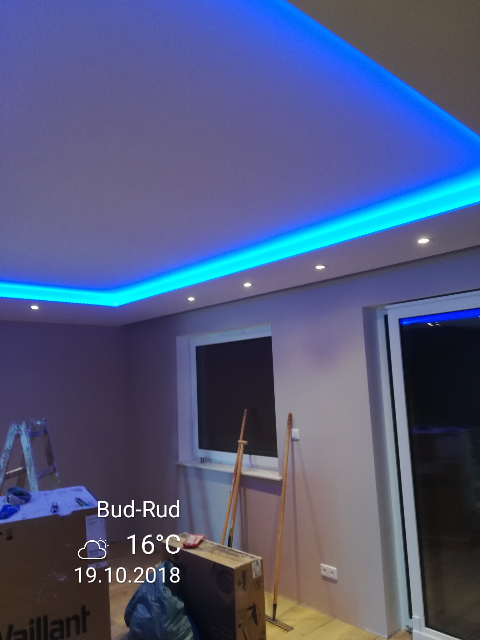 Pokój po remoncie z podwieszonym sufitem i oświetleniem LED w kolorze niebieskim, widoczne okno, drabina i kartony z wyposażeniem.