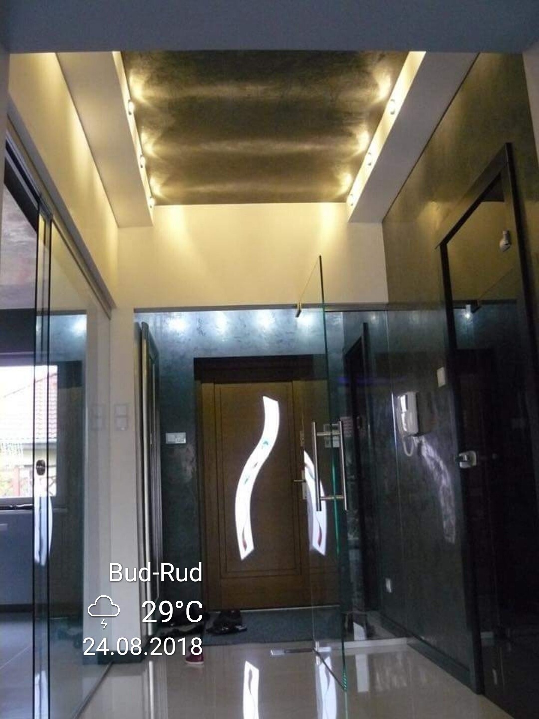 Wnętrze przedpokoju z podświetlanym sufitem w odcieniach szarości, nowoczesne drzwi z dekoracyjnym motywem, szklane drzwi przesuwne po lewej stronie, marmurowe ściany i podłoga.