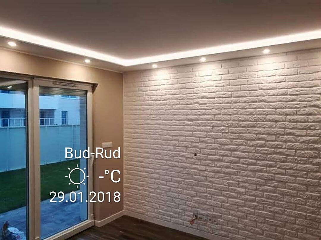 Wykończone wnętrze: ściana z białej cegły dekoracyjnej z oświetleniem LED podsufitowym, widoczne panele podłogowe i przesuwne drzwi balkonowe z widokiem na ogród.