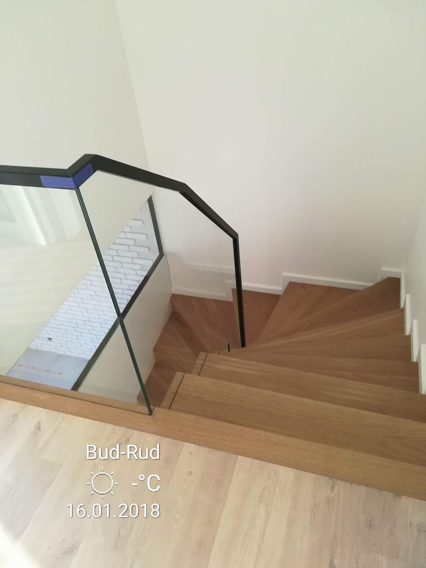 Drewniane schody spiralne z czarną, szklaną balustradą, widok z góry, białe ściany i podłoga w jasnym odcieniu drewna.