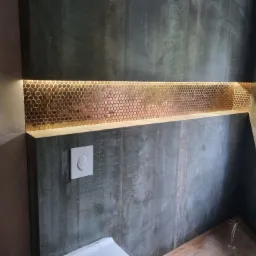 Nowoczesna łazienka z ciemnymi płytkami imitującymi beton, podświetlaną wnęką z mozaiką w odcieniach miedzi i białym przyciskiem spłuczki.