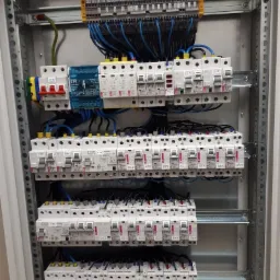 Rozdzielnica elektryczna z zamontowanymi bezpiecznikami i okablowaniem w szarym metalowym korpusie, widoczne oznaczenia na poszczególnych elementach.