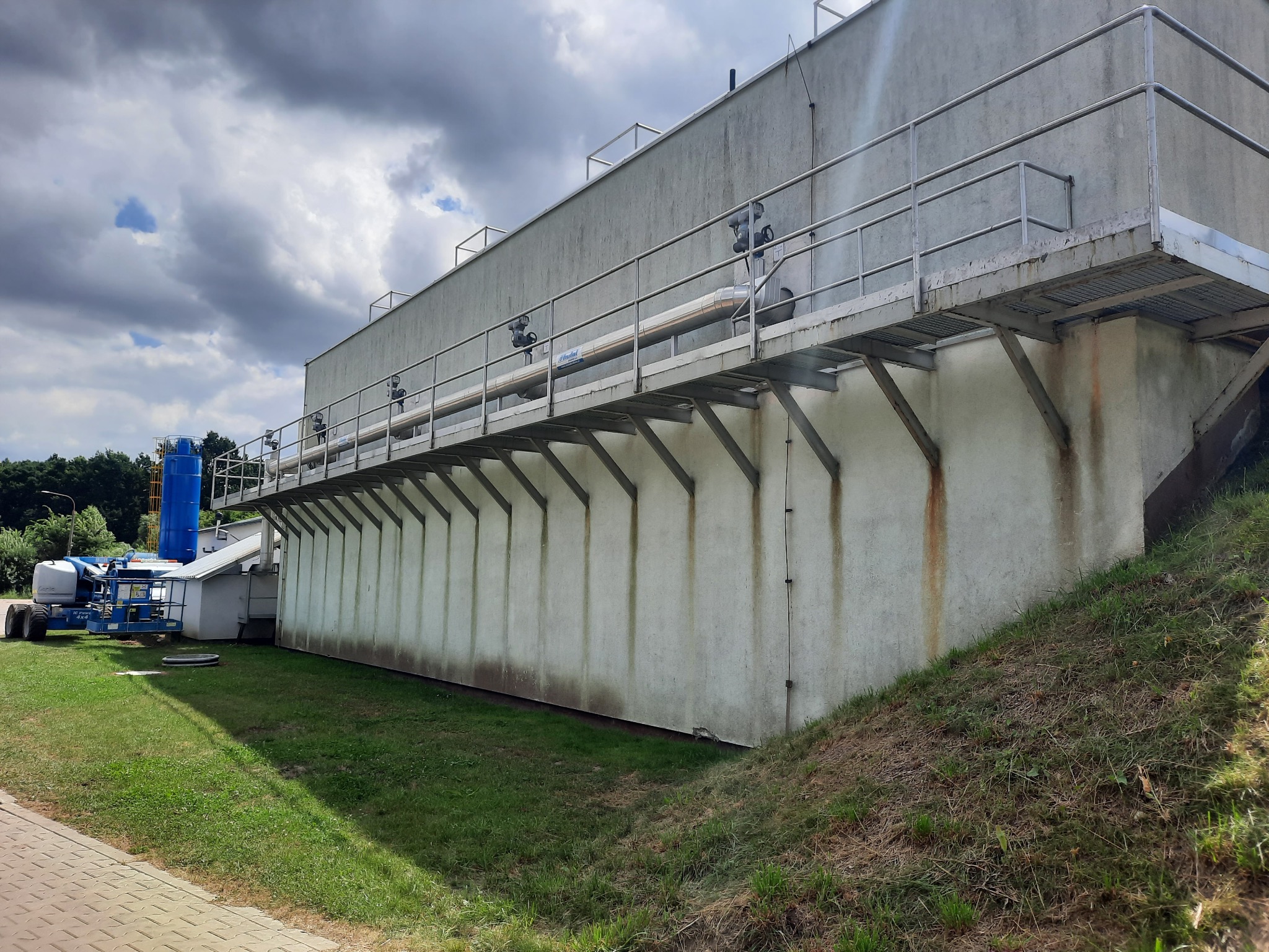 Zewnętrzna ściana budynku przemysłowego z metalową platformą i rurami, widoczna z poziomu gruntu, z podnośnikiem koszowym w tle.