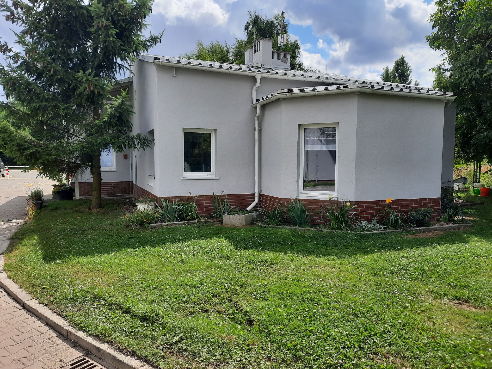 Parterowy budynek z szarą elewacją i ceglanym cokołem, widoczny z perspektywy ogrodu z drzewem iglastym po lewej stronie.