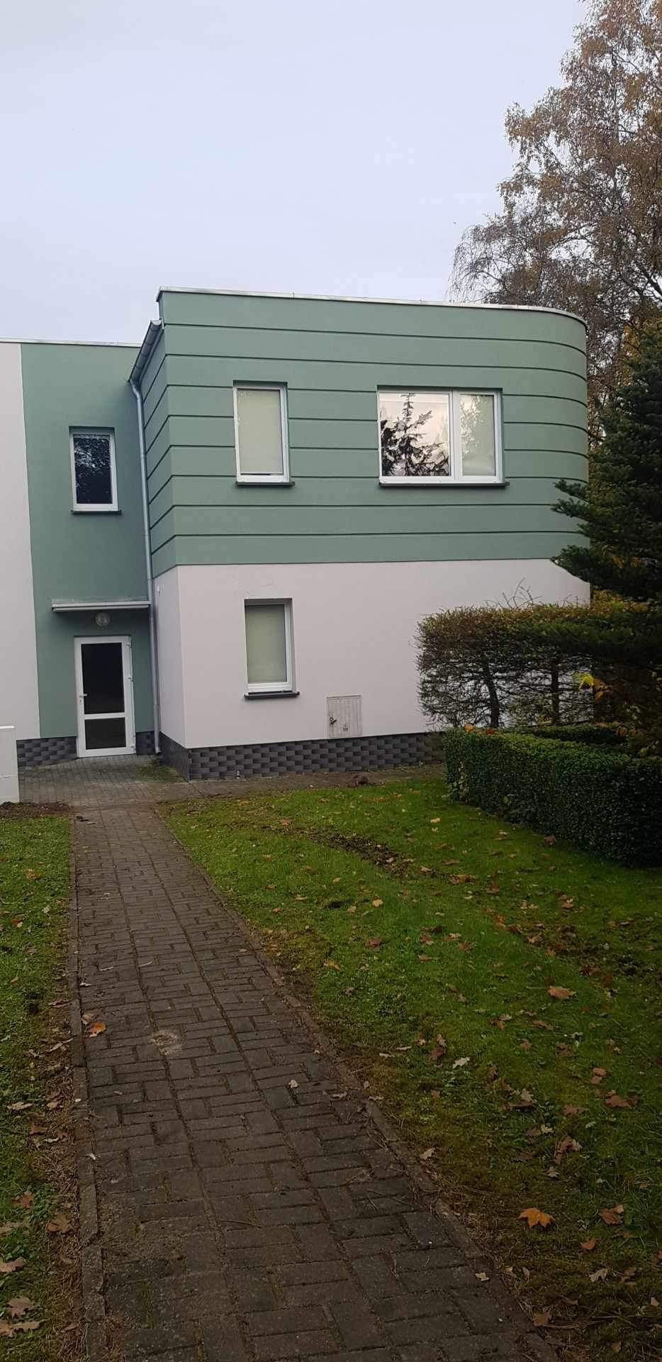 Dwukondygnacyjny dom z elewacją w odcieniach zieleni i bieli, z zaokrąglonym narożnikiem i widoczną ścieżką z kostki brukowej.