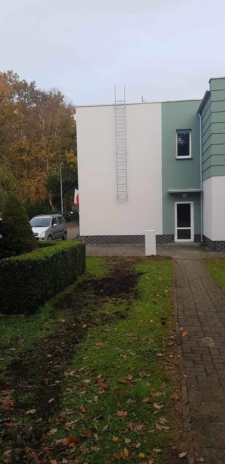 Dwukondygnacyjny budynek z biało-zieloną elewacją, z przymocowaną na stałe metalową drabinką na dachu, widok z poziomu trawnika z opadłymi liśćmi.