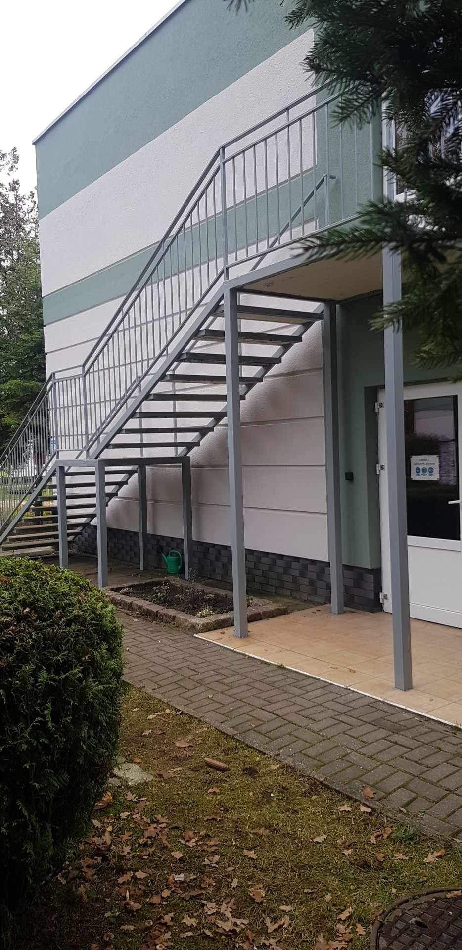 Zewnętrzne schody stalowe z szarymi barierkami prowadzące do budynku z elewacją w pasy, zielona konewka pod schodami.