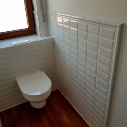 Wykończona łazienka z białymi, wypukłymi płytkami imitującymi cegiełki ułożonymi do połowy ściany, brązową drewnopodobną podłogą i wiszącą toaletą z białej ceramiki.