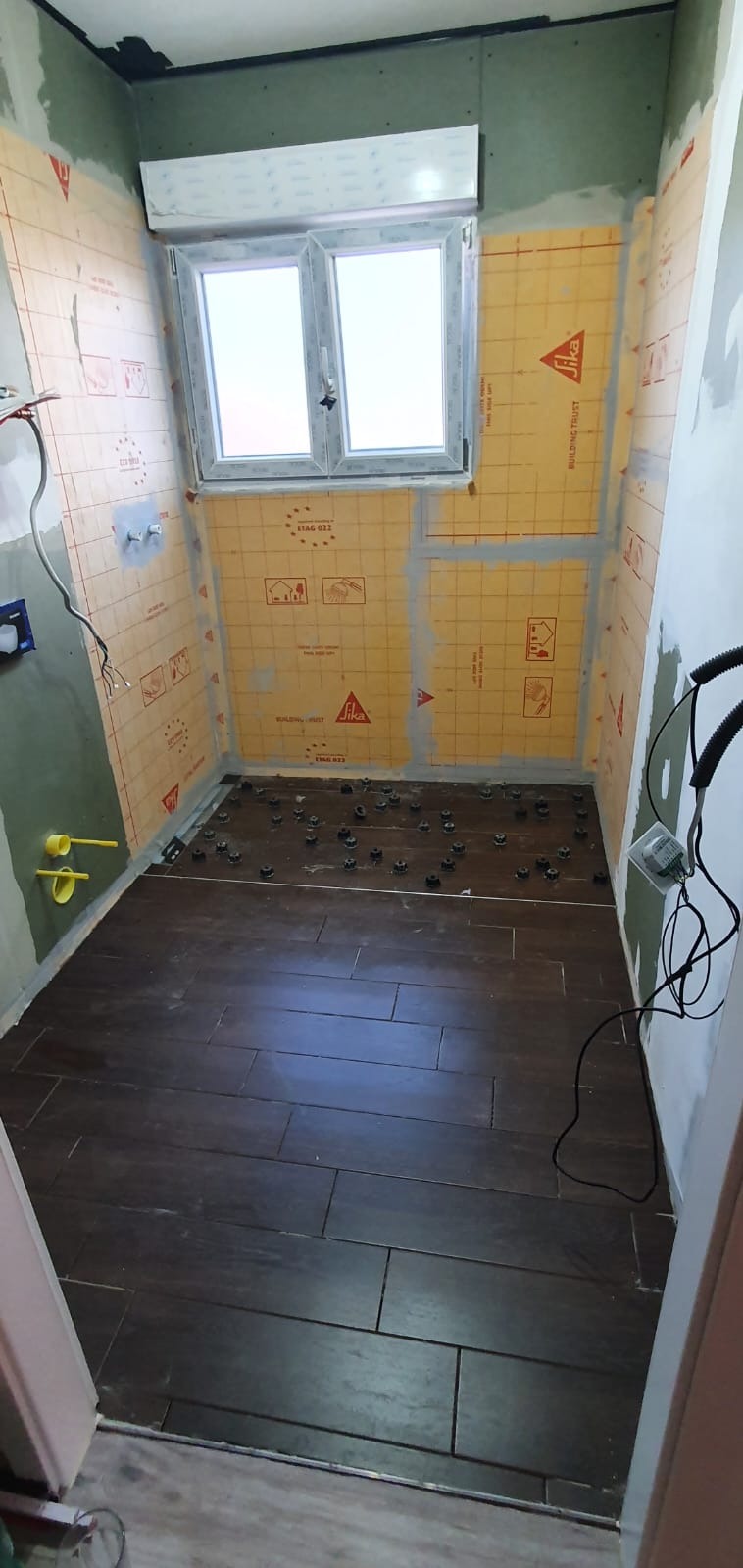 Wnętrze łazienki w trakcie prac remontowych: brązowe płytki ułożone na podłodze, ściany przygotowane z żółtą membraną izolacyjną z nadrukiem, widoczne elementy instalacji wodnej i okno.