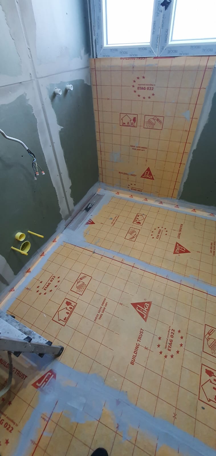 Przygotowanie podłoża w łazience z użyciem maty izolacyjnej Sika Building Trust ETAG 022, widoczne niedociągnięcia w postaci szpachli, surowe ściany i wystające kable.