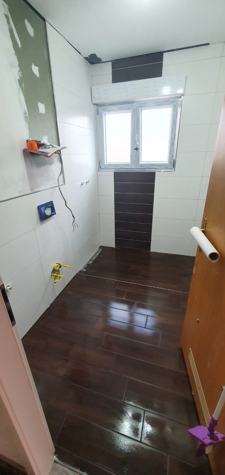 Łazienka w trakcie remontu: ułożona ciemnobrązowa podłoga z płytek imitujących drewno, białe płytki na ścianach z fragmentem ciemnobrązowych płytek dekoracyjnych pod oknem, widoczne instalacje...